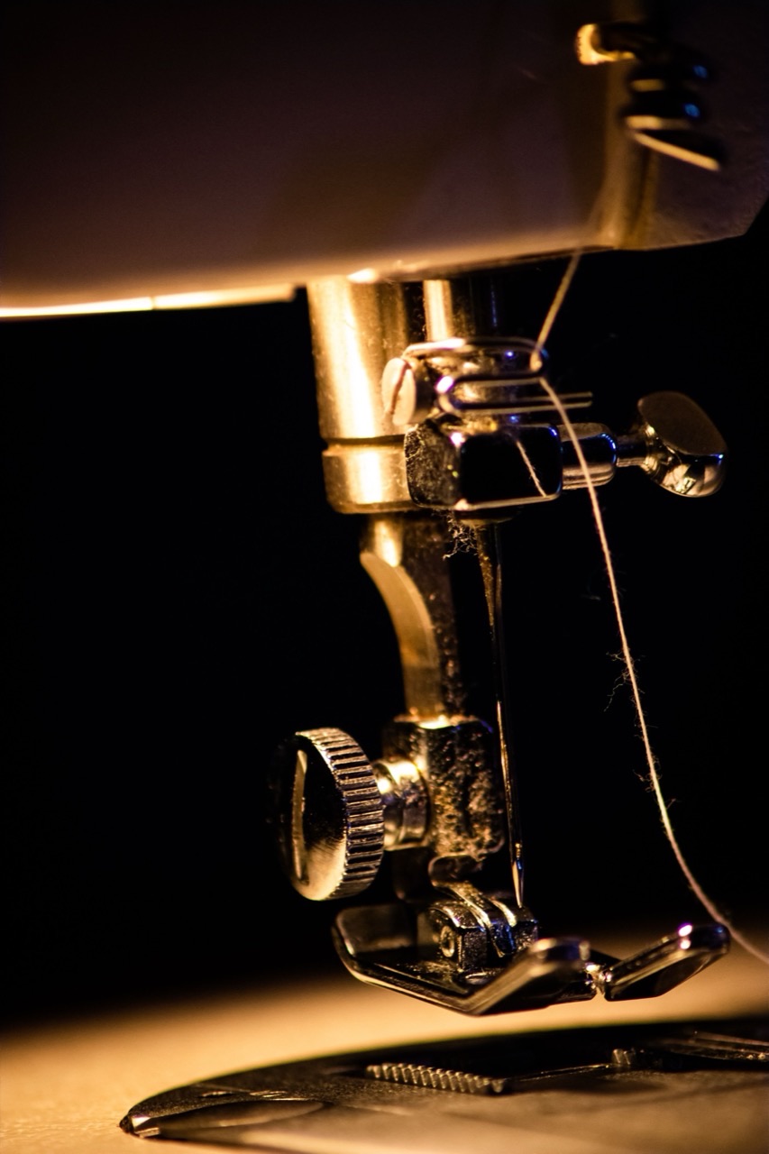 Close-up of an embroidery machine needle and presser foot, warmly lit against a dark background with thread running down to the plate.