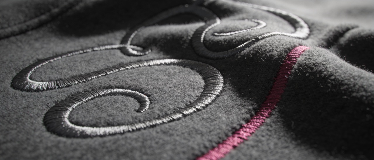 Extreme macro of dimensional satin-stitch embroidery across charcoal fleece, thread catching a low raking light.