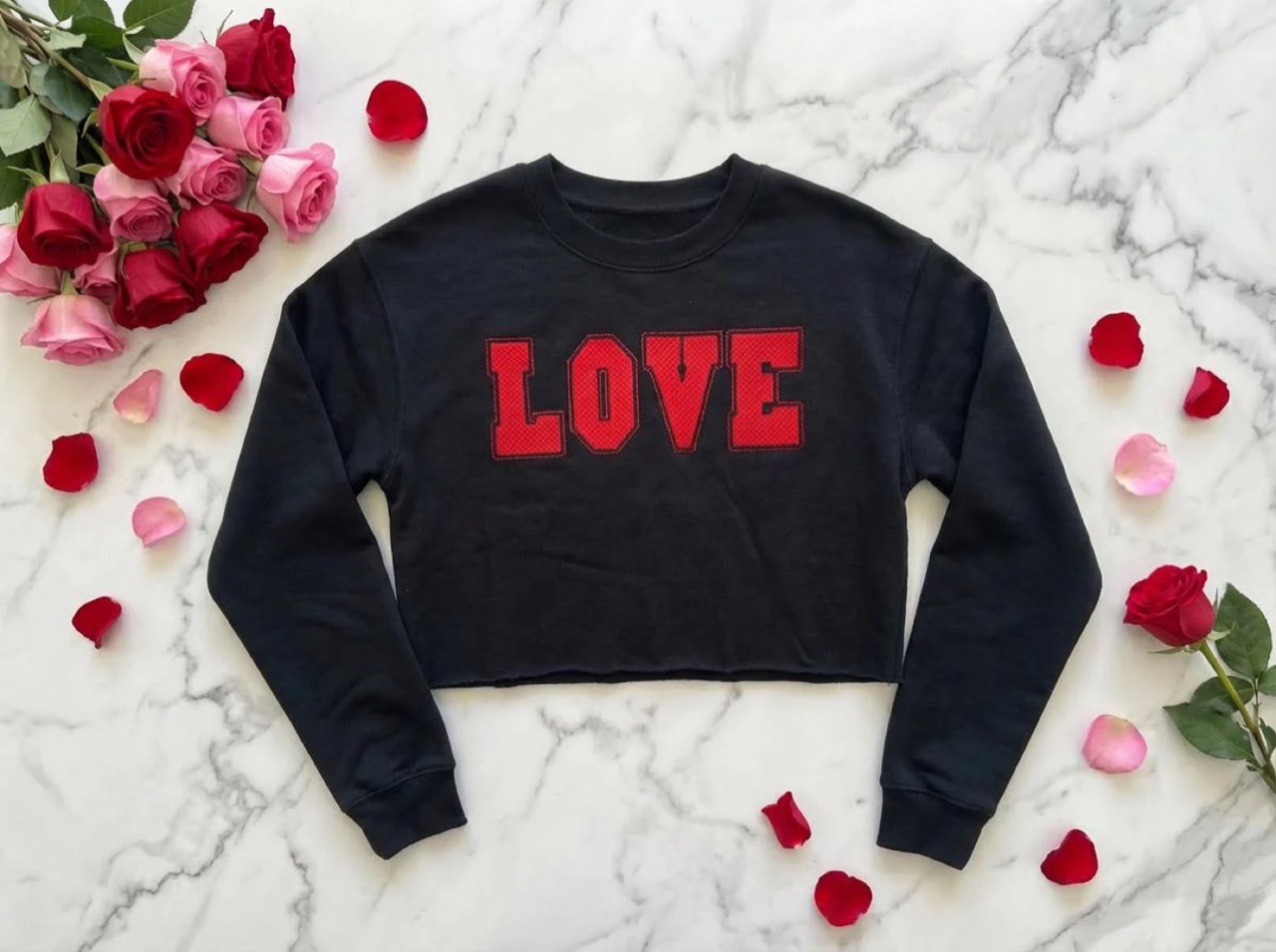 A black cropped crewneck sweatshirt laid on marble, with LOVE across the chest in red appliqué lettering, surrounded by roses.