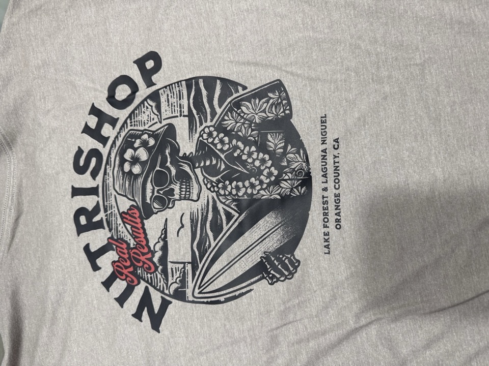 A heather grey tee printed with a circular surf and skull illustration reading Nutrishop, Real Results, Lake Forest and Laguna Niguel, Orange County, California.