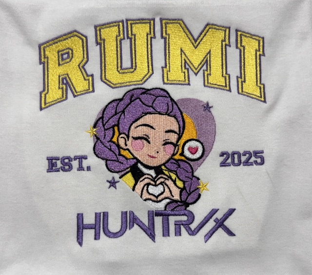 Dense multi-colour embroidery on white fabric: RUMI arched in gold above an illustrated character with purple braids, and HUNTRIX beneath.